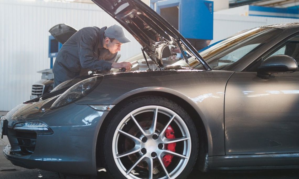 Male mechanic in a gray uniform is inspecting the hood of a gray Porsche sports car in an auto garage shop.