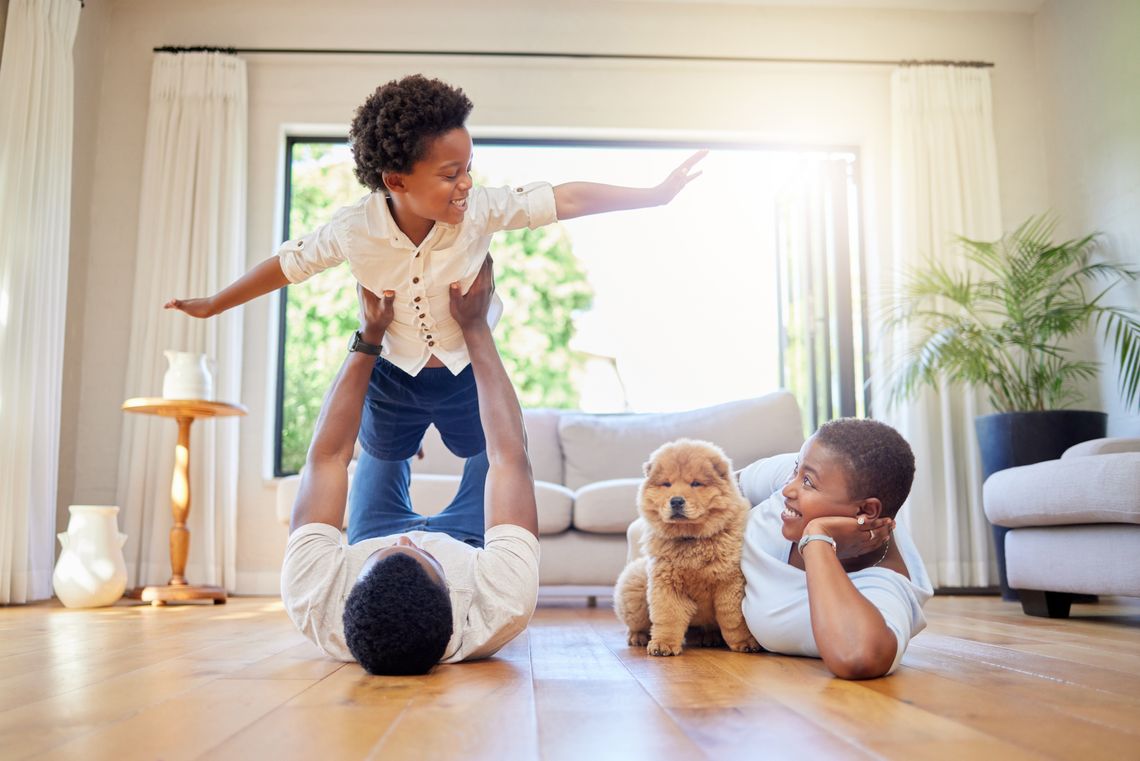 How to Choose Floors That Fit Your Family’s Lifestyle A dad and mother lying on a living room hardwood floor with their child raised on legs and a small dog beside them.