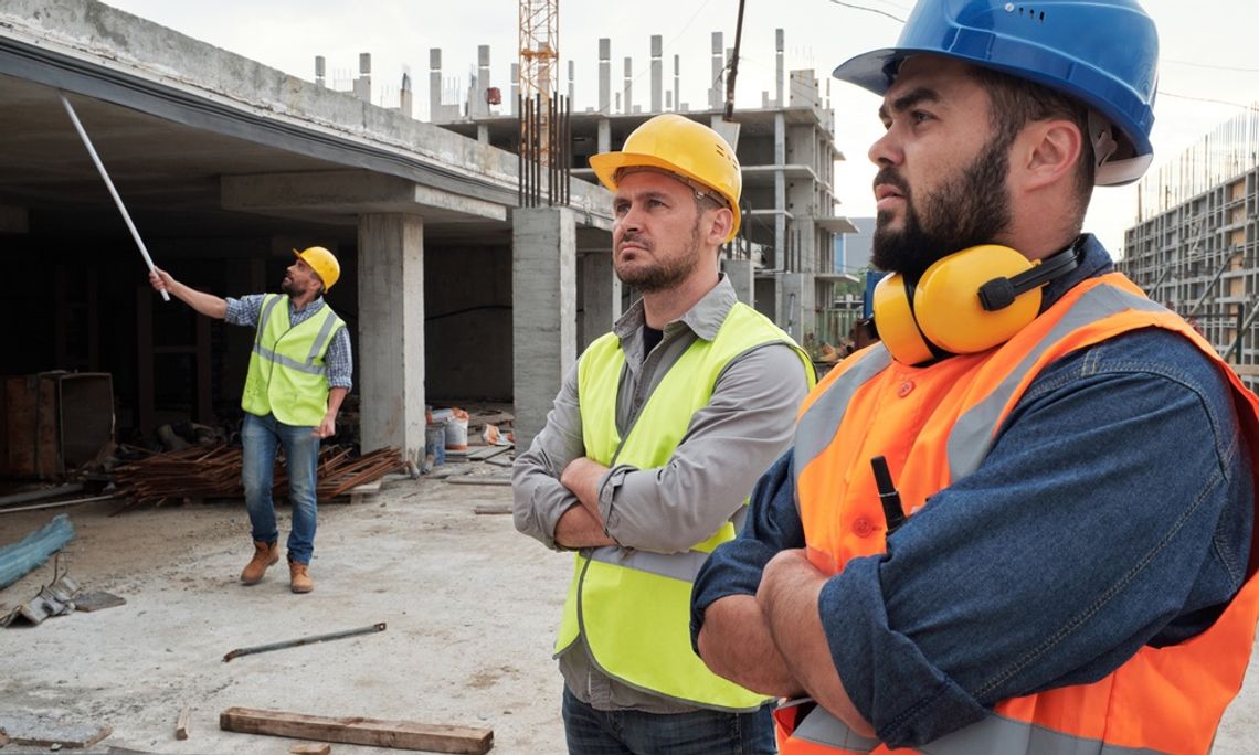 Three workers in safety vests and hard hats on a jobsite. Two are observing while the other checks part of the construction.
