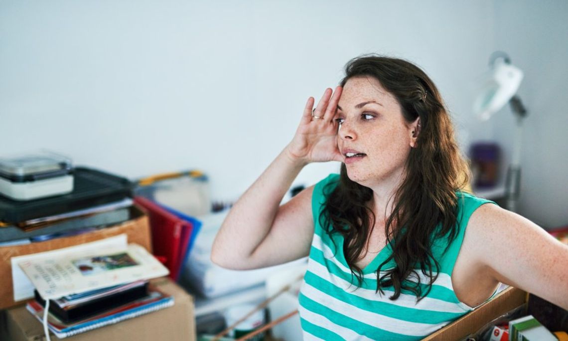 A woman wearing a green and white shirt holds her head while looking at piles of clutter. She's also holding a box of items.