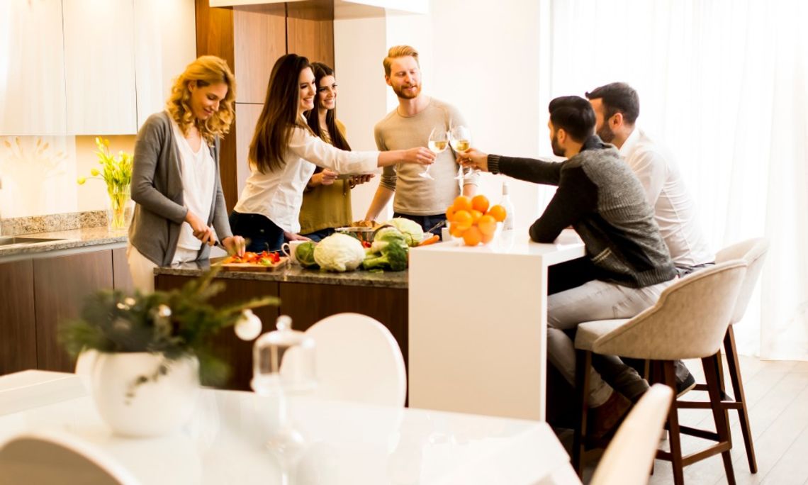 How to Create a Kitchen That Works for Entertaining Four people raising glasses of white wine over a kitchen table with plates of food and blurred lights behind them.