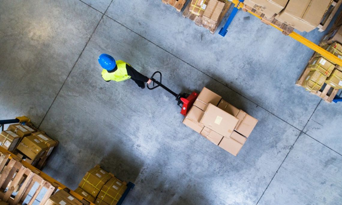 How To Create a Safer, More Organized Warehouse An aerial view of a warehouse employee in PPE gear rolling a dolly behind them. The dolly has boxes on it.