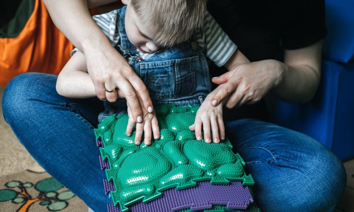 A young boy sits in the lap of a woman while being guided to participate in an activity with a sensory mat.