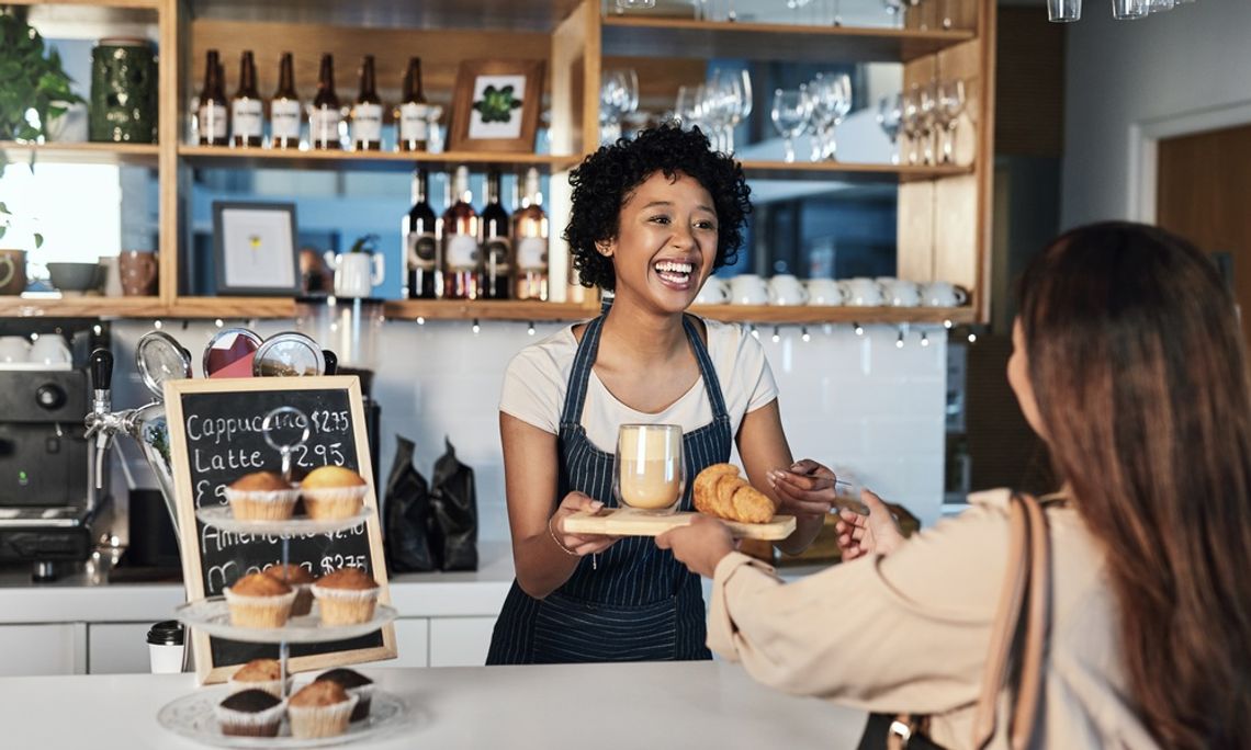 A smiling female barista hands a latte and pastry to a female customer at a cozy coffee shop counter.