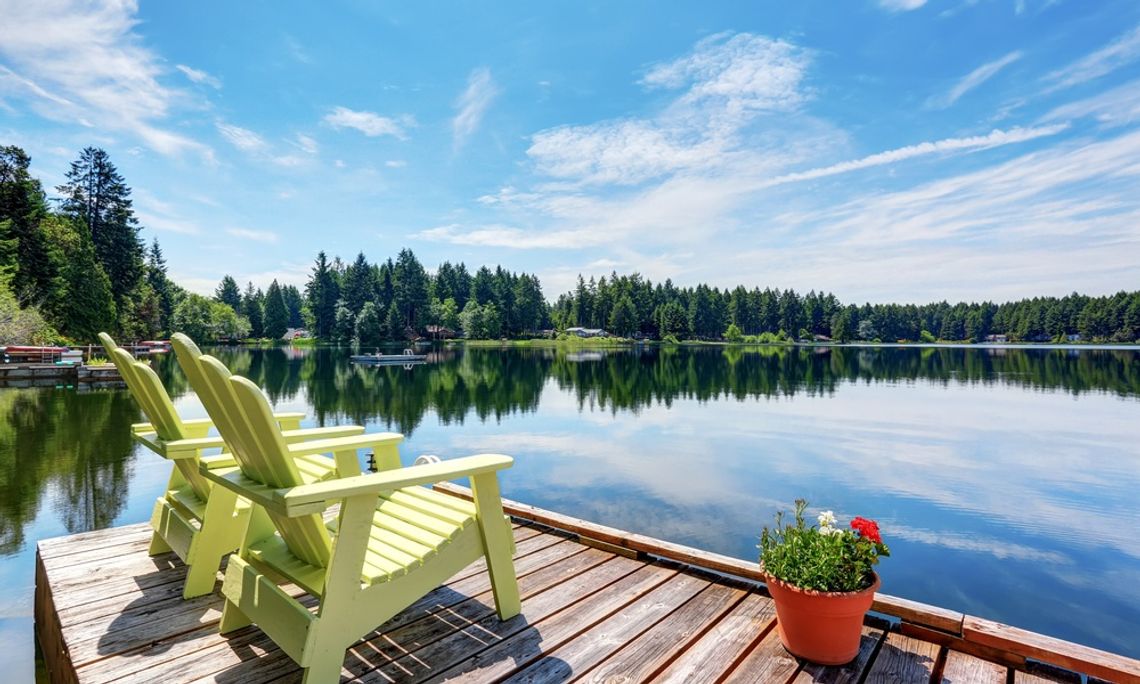 A wooden pier with two yellow Adirondack chairs and a potted plant, overlooking a lake surrounded by trees.
