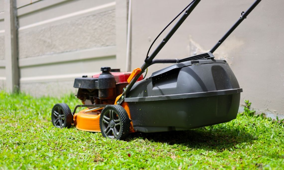 An orange and black push lawn mower with a rear grass collection bag resting on green grass beside a concrete wall.