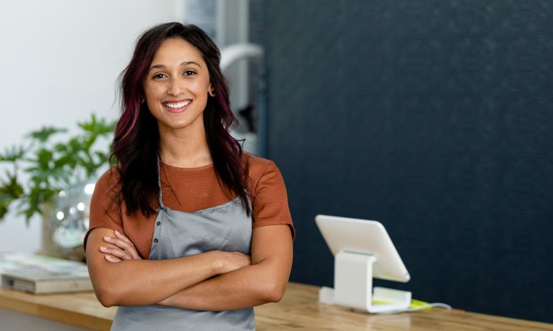 A smiling woman wearing an orange shirt and a gray apron stands in front of a reception area with crossed arms.