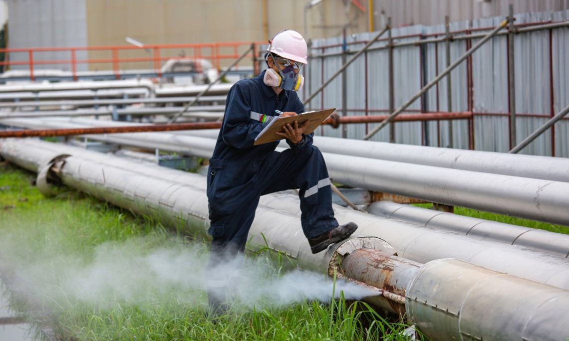 A male worker in full PPE writes on a clipboard and stands with his foot on a gas line that is emitting a stream of steam.