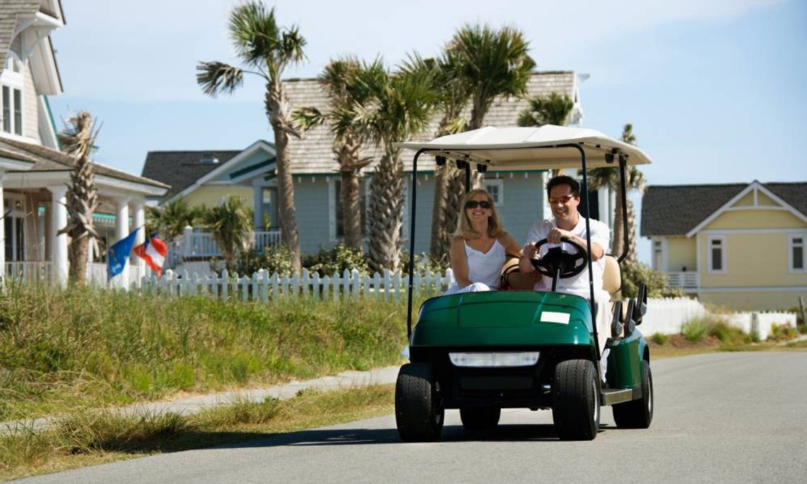 Two people in a golf cart on the road. There are houses, fences, and palm trees in the distance.