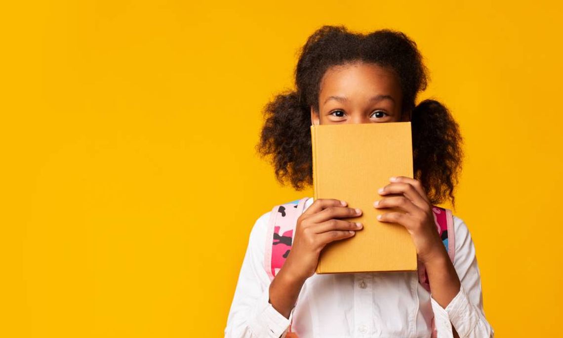 A young girl holds a yellow book in front of the lower half of her face in front of a yellow background.