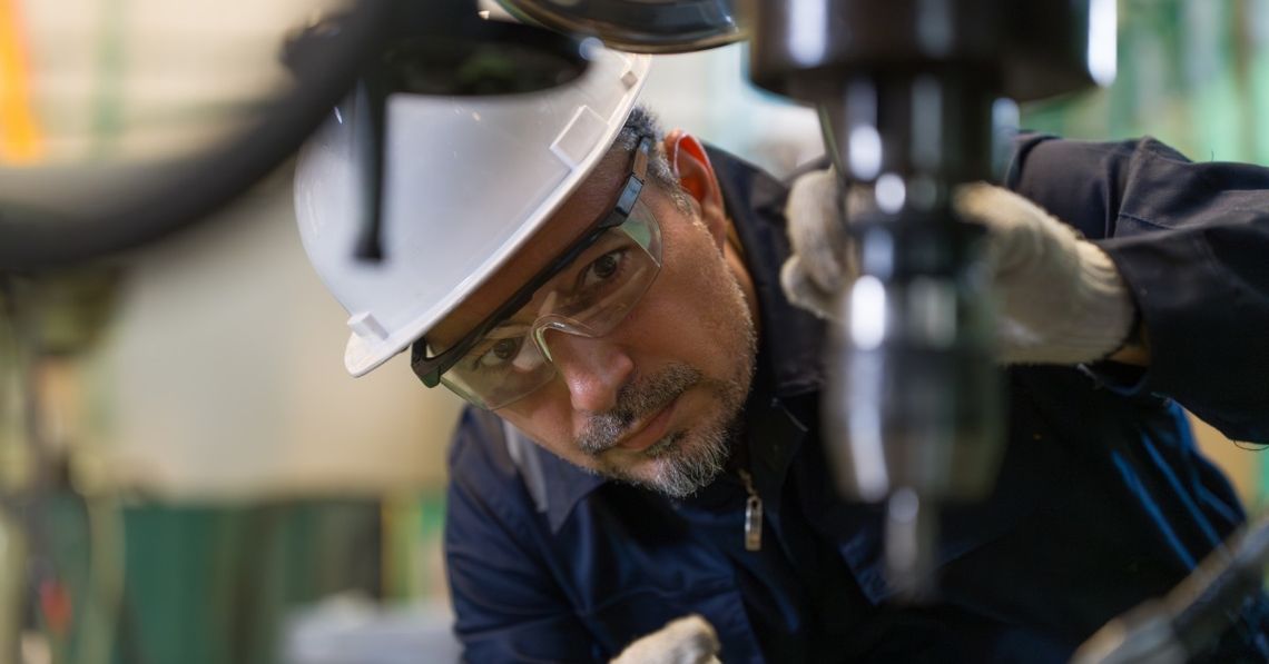 A close-up of a man in a hard hat and safety goggles with gloves crouching to inspect an industrial drill machine.