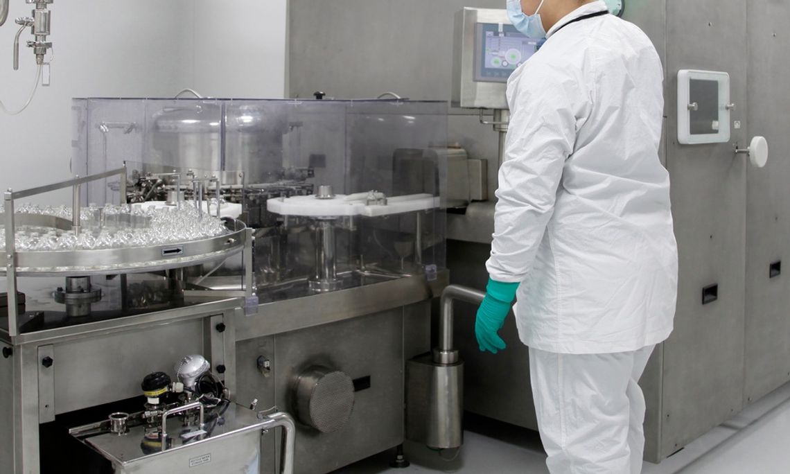A rear view of a person in a lab coat, protective gloves, and a face mask standing in front of a drug production line.