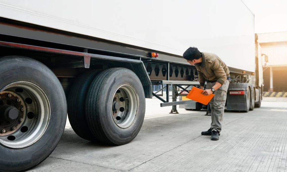 How to Keep a Work Semi-Truck From Wearing Out Early A man holding an orange clipboard while bending down to inspect the wheels and underside of a large white truck.
