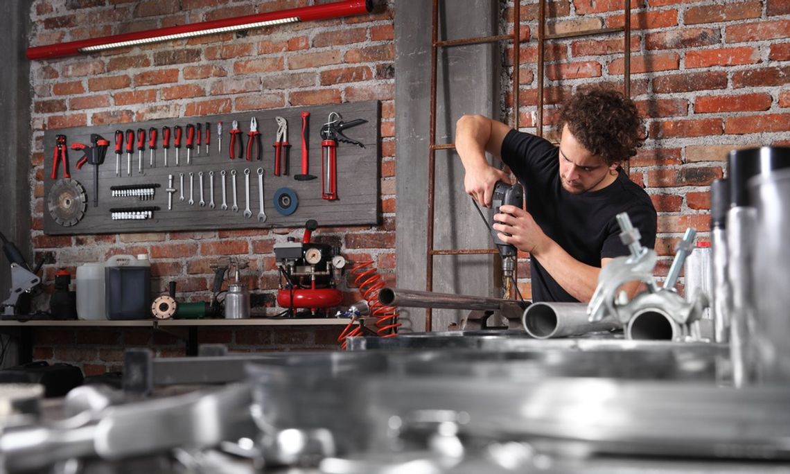 A man using a drill on a metal tube in his workshop. There is a lot of metal lying around, and some tools hung up on the wall.