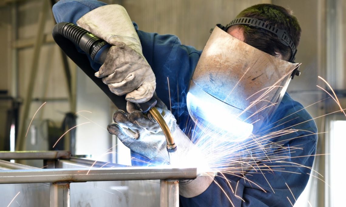 How to Learn Welding Without Breaking the Bank A person wearing a welding helmet, blue work jacket, and protective gloves welding a metal structure as sparks fly.