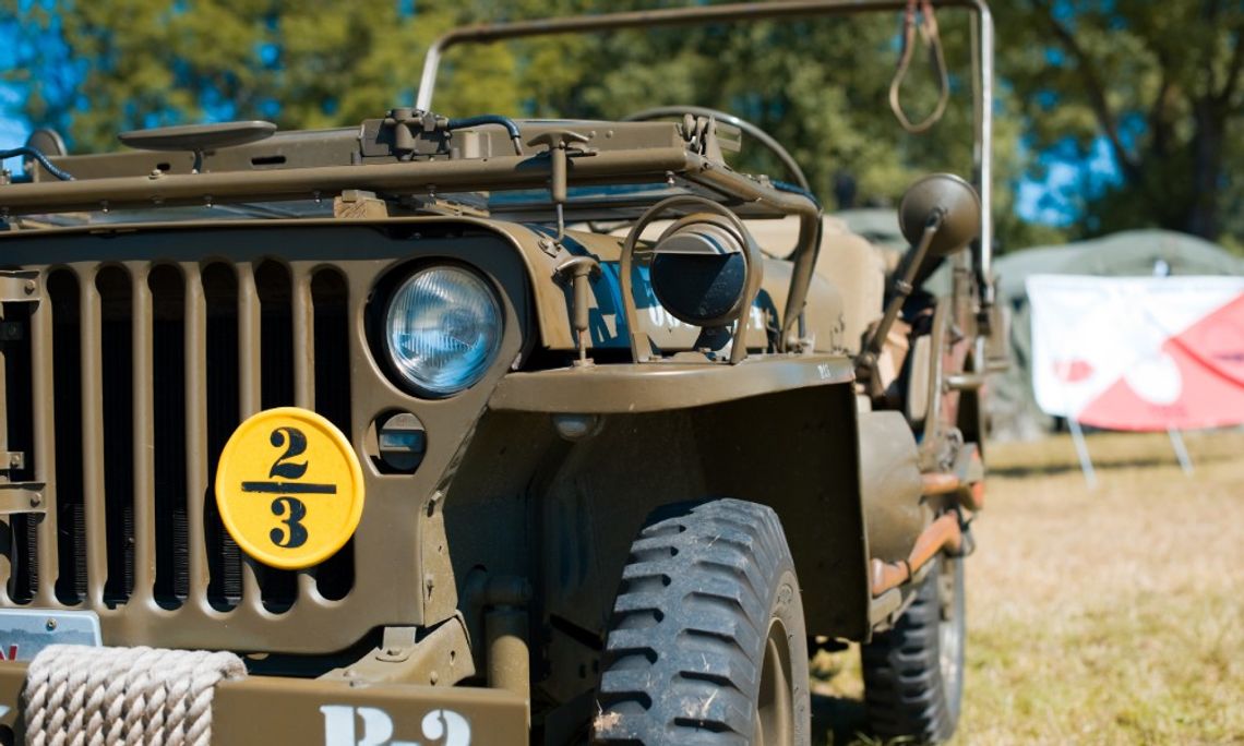 Half of the front part of an olive green jeep with a yellow badge on the grill. The jeep sits on grass near some tents.