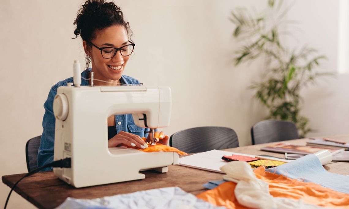 How To Make Sewing Your New Mindful Hobby A young woman using a sewing machine and smiling at her table. The table is covered in bits of other fabric.
