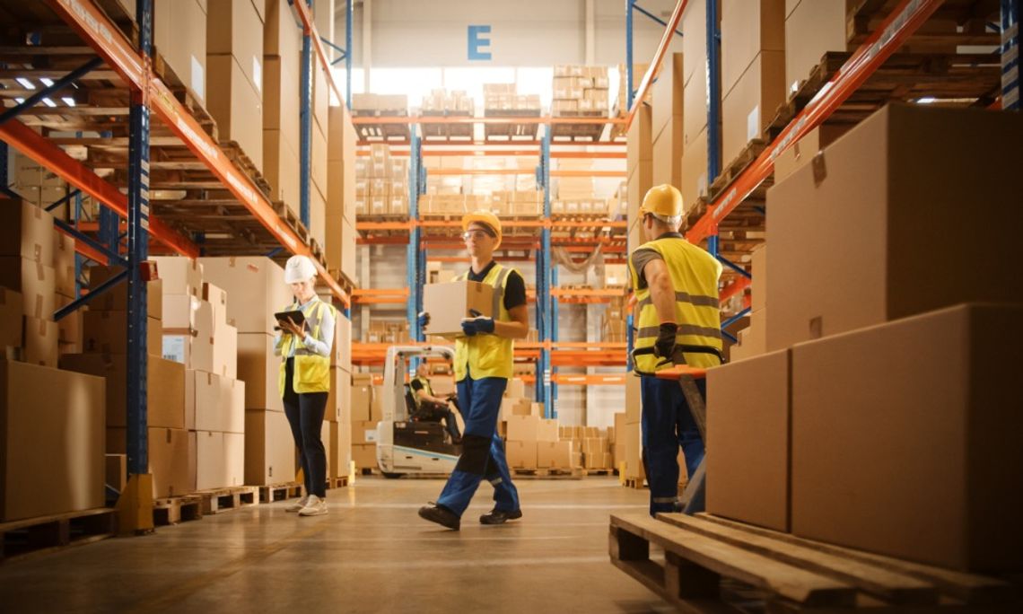 Three warehouse workers wearing hard hats and safety vests handling boxes and checking inventory between storage racks.