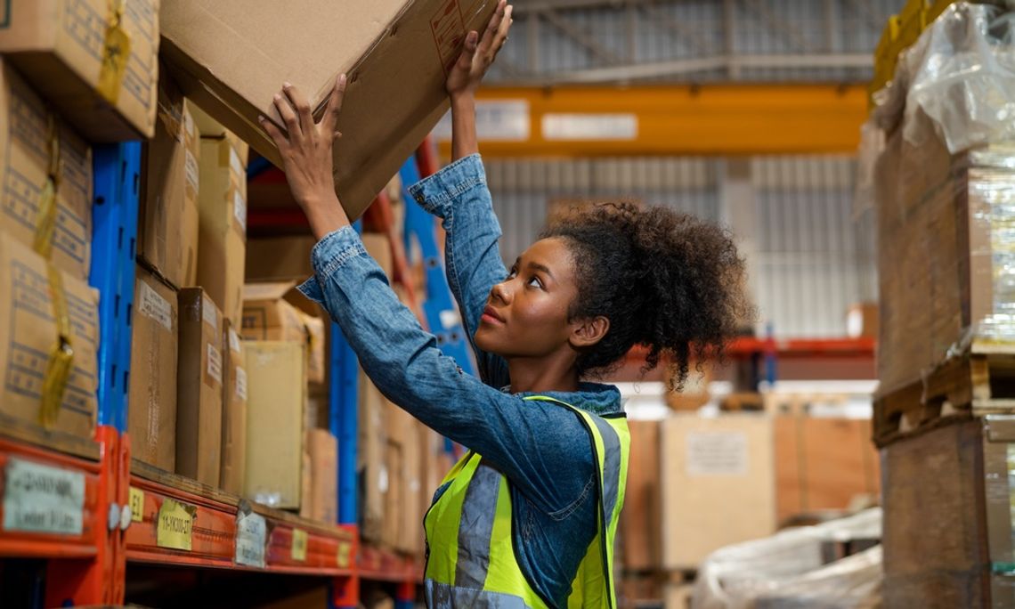 A warehouse worker wearing a safety vest reaches up to place a box on a high shelf among stacked packages.