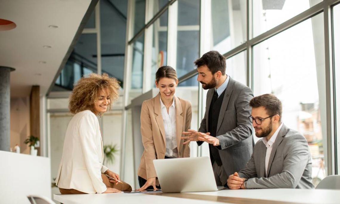 Four employees looking at a single laptop enthusiastically in their office next to a set of large windows.