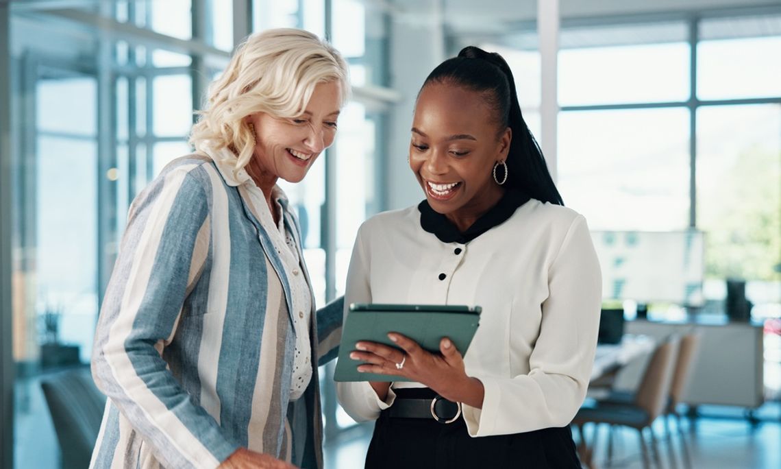 Two happy women business partners are looking at a tablet happily and pointing at the screen outside a conference room.