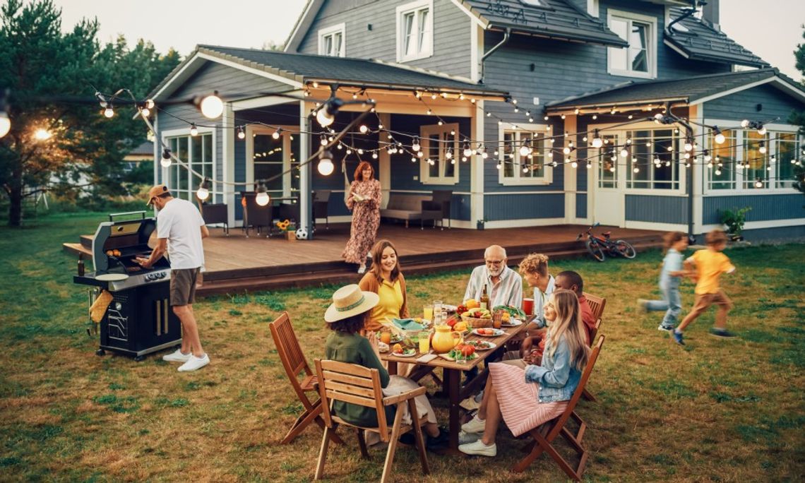 A family is sitting at an outdoor table eating a meal. A man is using the grill and children are running around the backyard.