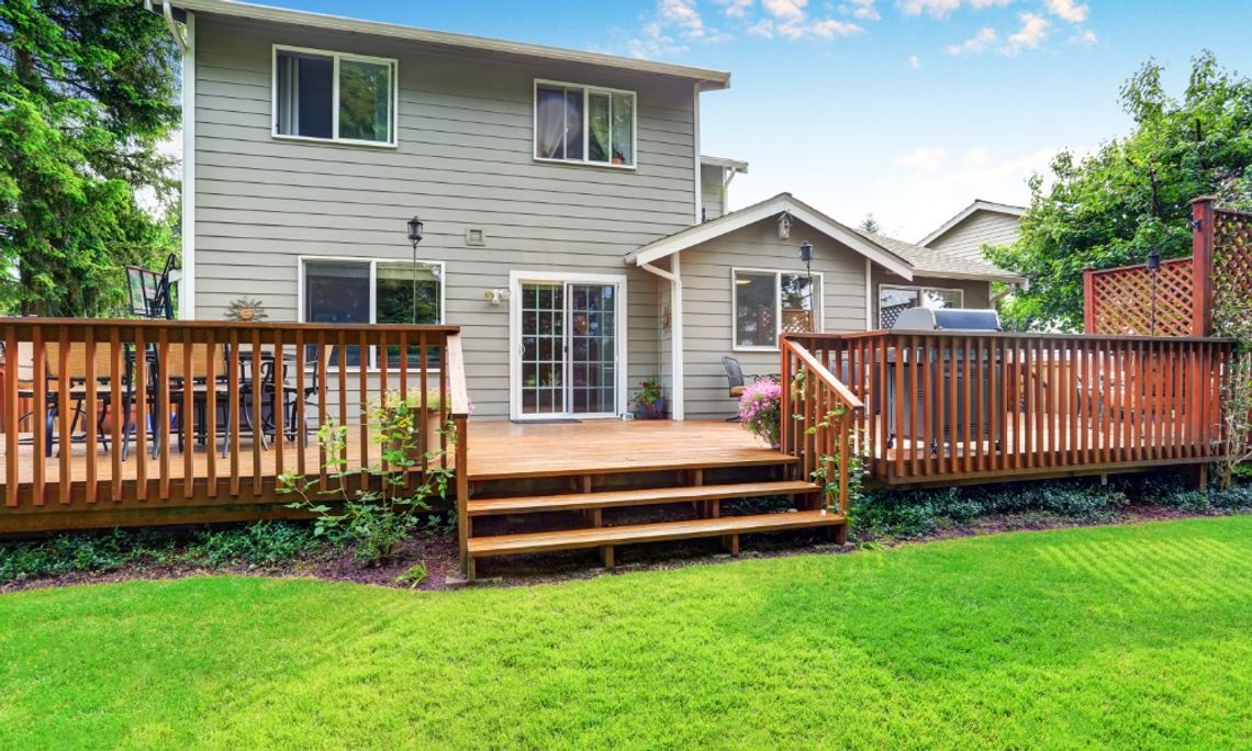 How To Modernize a Deck Without Rebuilding It A wooden backyard deck with railings, stairs, outdoor seating, and a grill overlooking a green lawn behind a two-story home.