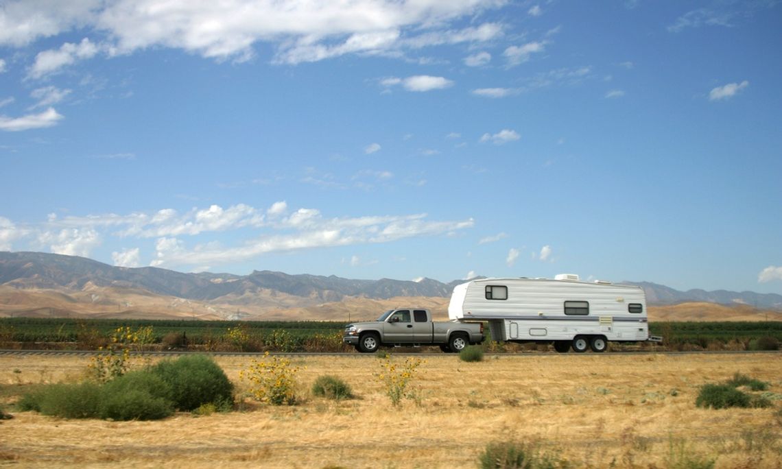A grey pickup truck towing a large white camper trailer on an empty road with a mountain ridge in the background.