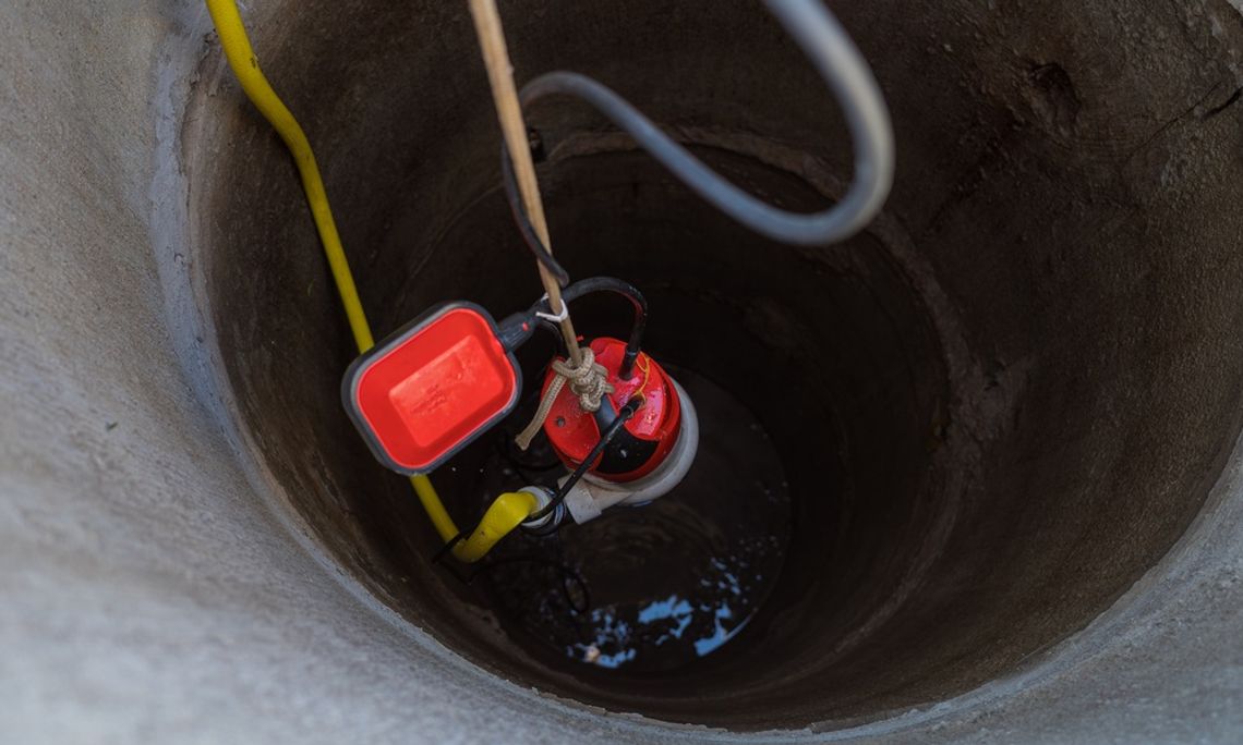 A red pump being lowered into a concrete well with ropes, a yellow hose, black cable, gray looped pipe, and water below.