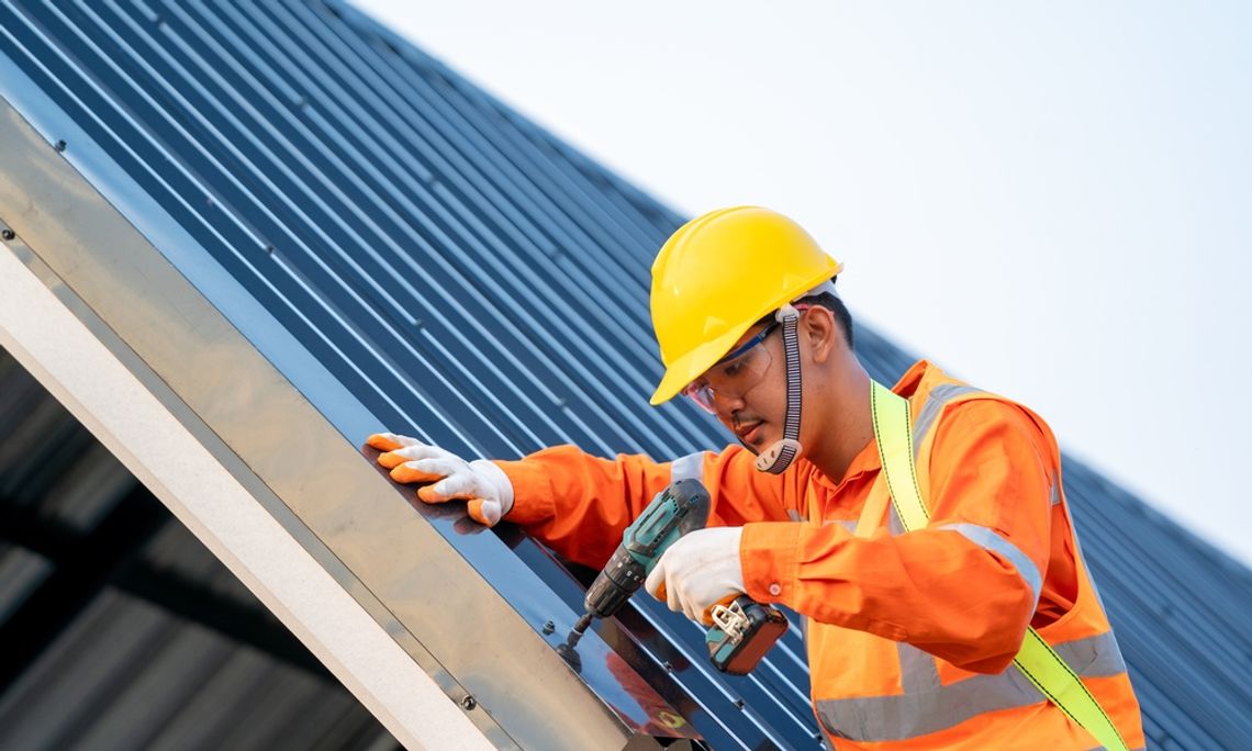 A worker wearing a hard hat installing roof panels on a metal roof frame with blue sky in the background.