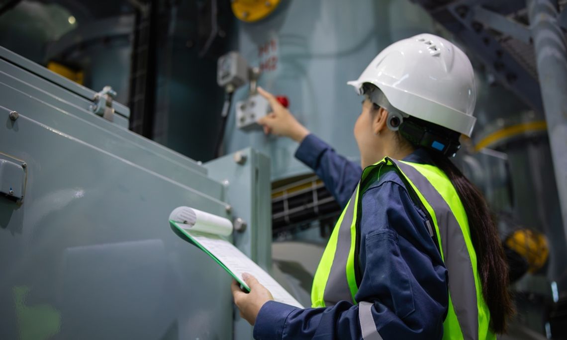 How To Prepare for a Commercial Energy Audit A woman in a white hard hat and green safety vest holds a green clipboard and points toward large equipment.