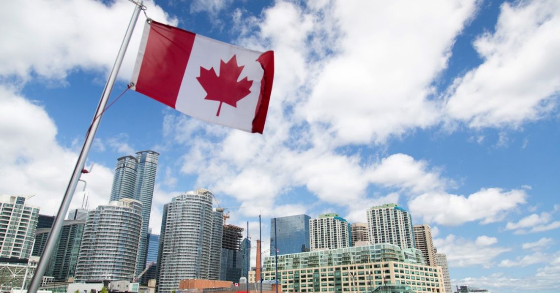 How To Prepare for a Road Trip to Canada Canadian flag waving above Toronto’s waterfront skyline beneath a blue sky filled with large white clouds.