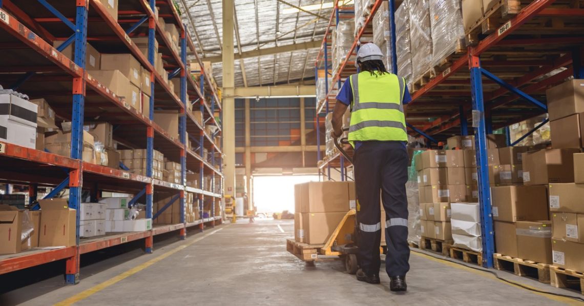 How to Prepare Storage Systems for Inventory Changes A worker wearing a hard hat and safety vest pulling a pallet truck with boxes through a warehouse aisle.