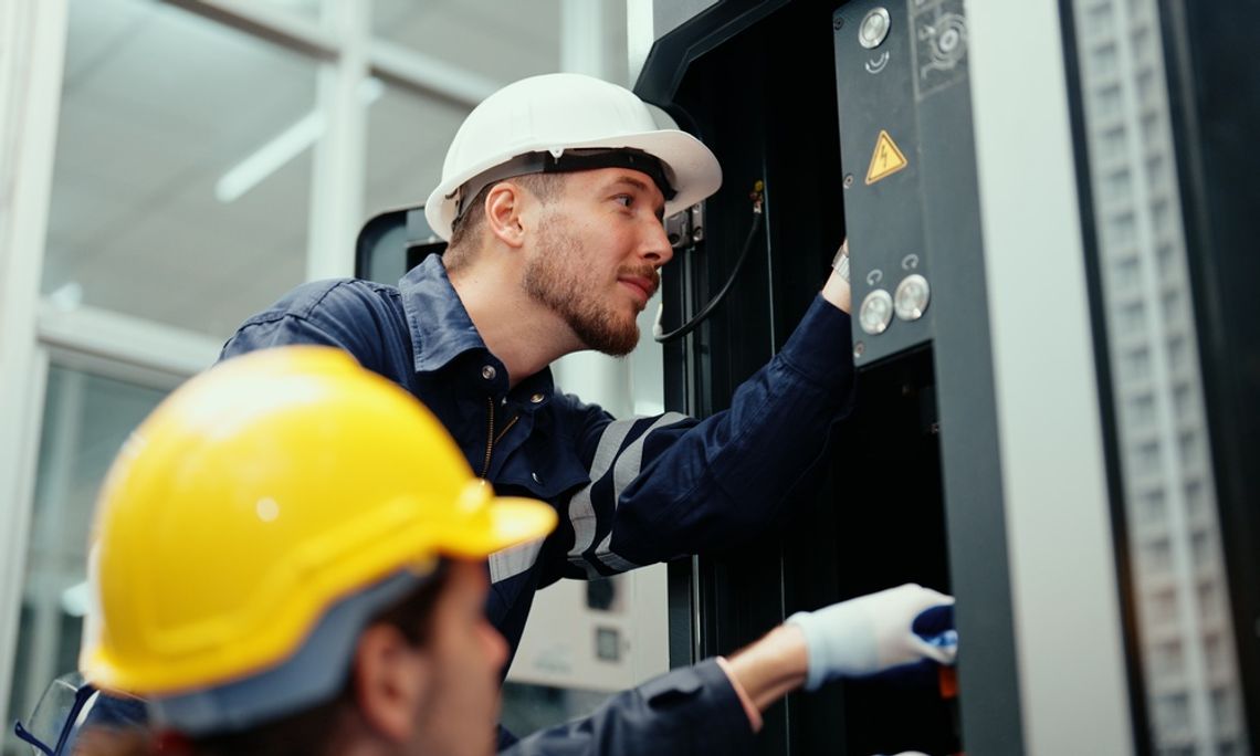 Two engineers wearing hard hats in a brightly lit factory. Both employees make repairs to a large CNC machine.