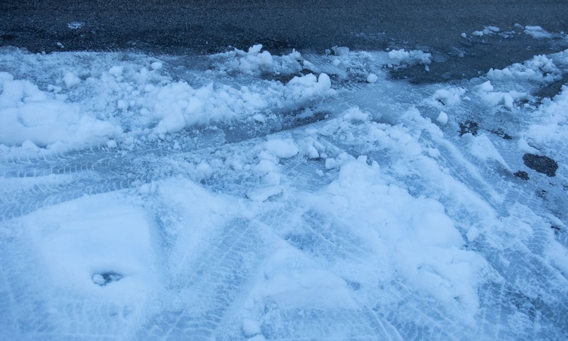 A thin layer of slush covering dark asphalt pavement, marked by tire tracks and some rough, broken-up patches.