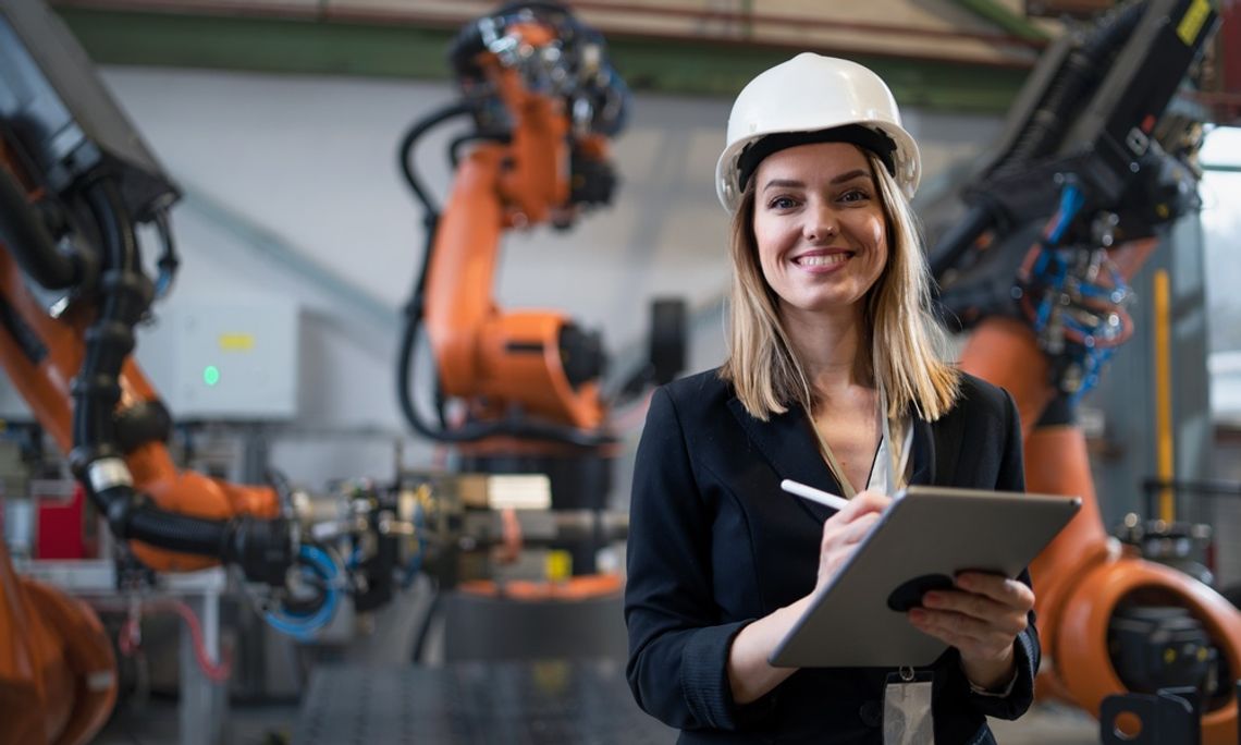 A woman in business clothing and a hard hat stands in front of three manufacturing robots. She holds a tablet in her hand.