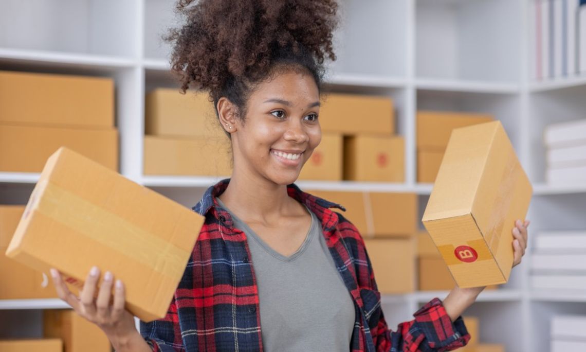 A young Black professional businesswoman is smiling while holding two cardboard boxes in a shipping center.