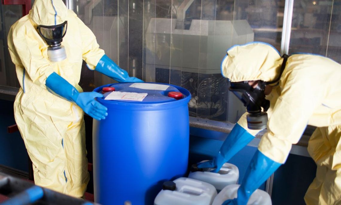 Two people wearing identical protective suits, gloves, and facial coverings stand with chemical storage containers.
