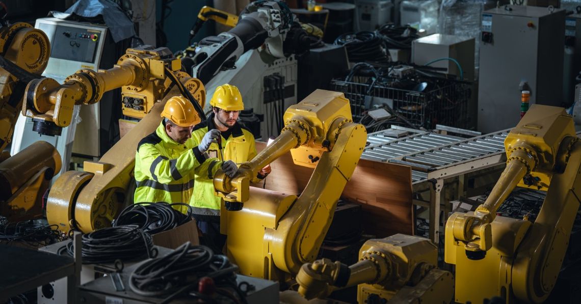 Two engineers wearing yellow hard hats and high-visibility vests inspect a long, yellow robotic arm in a factory.