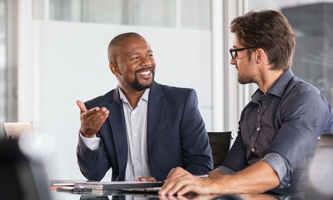 How To Turn Vision Into Actionable Business Plans Two men are smiling and talking to one another in a formal business setting. They're both wearing suits.