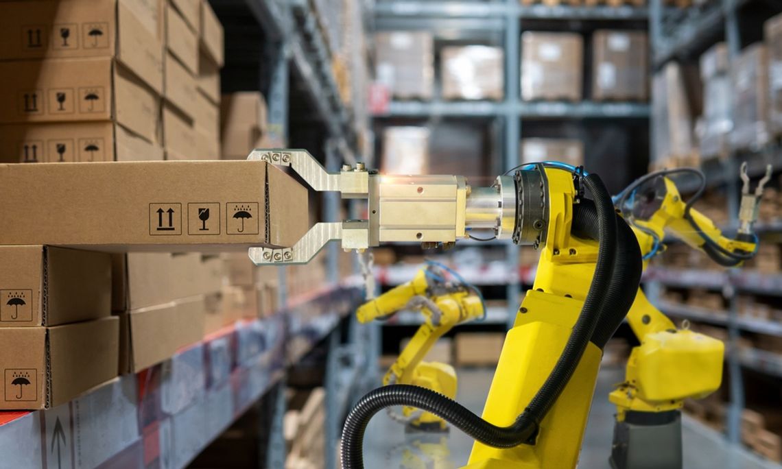 How Warehouse Automation Improves Material Handling A yellow robotic arm grips a cardboard box beside stacked boxes and shelves in the aisle of a warehouse.