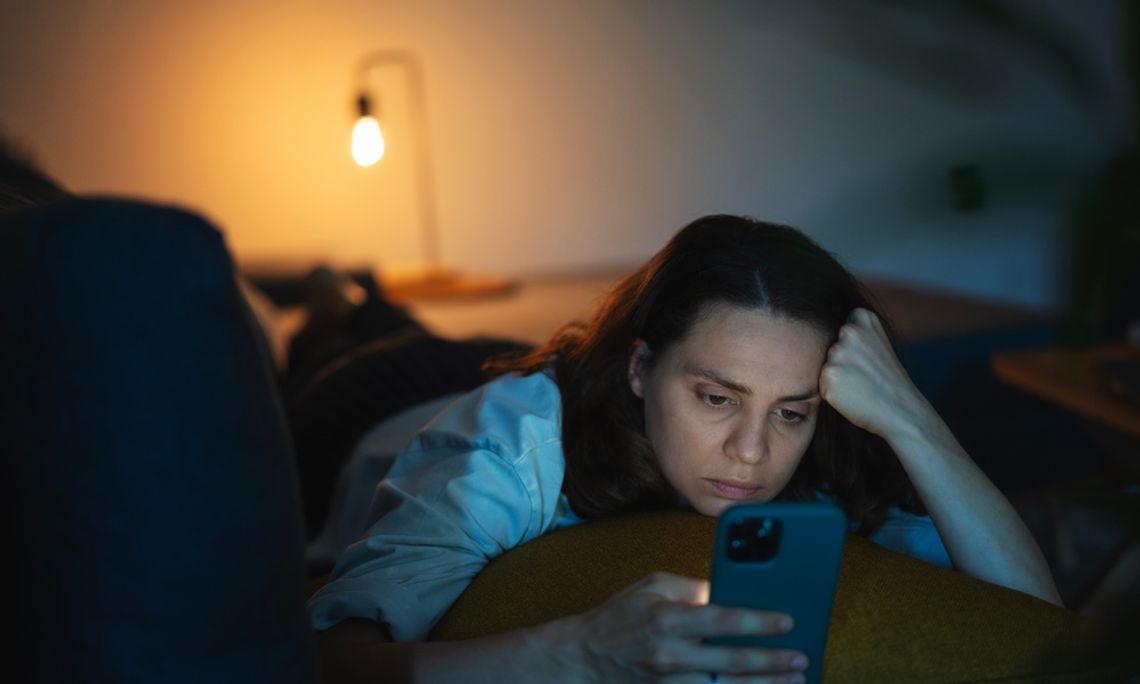How Workplace Burnout Can Affect After-Hours Life In a dim room, a woman lies on her stomach on a couch. She props her head up with her arm as she looks at her cellphone.