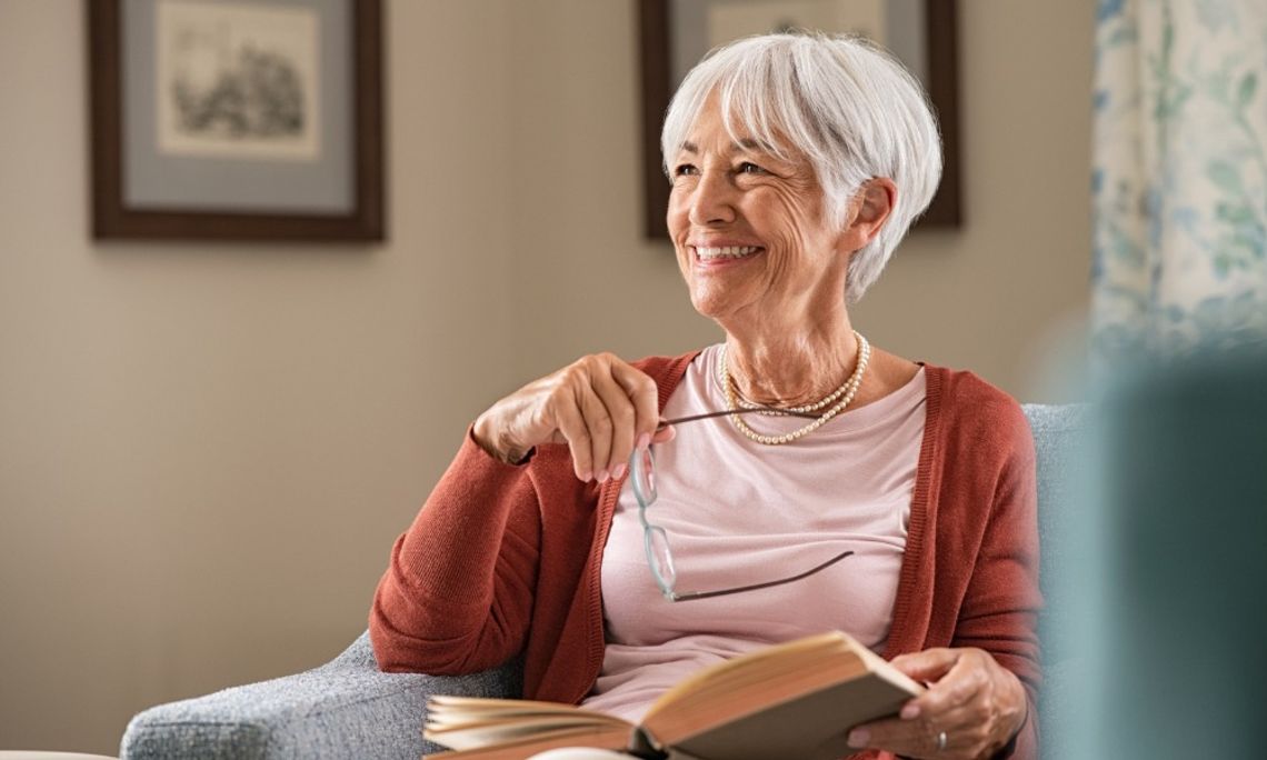 A smiling older woman with short gray hair sits in an armchair holding a book and glasses, relaxing in a cozy living room.