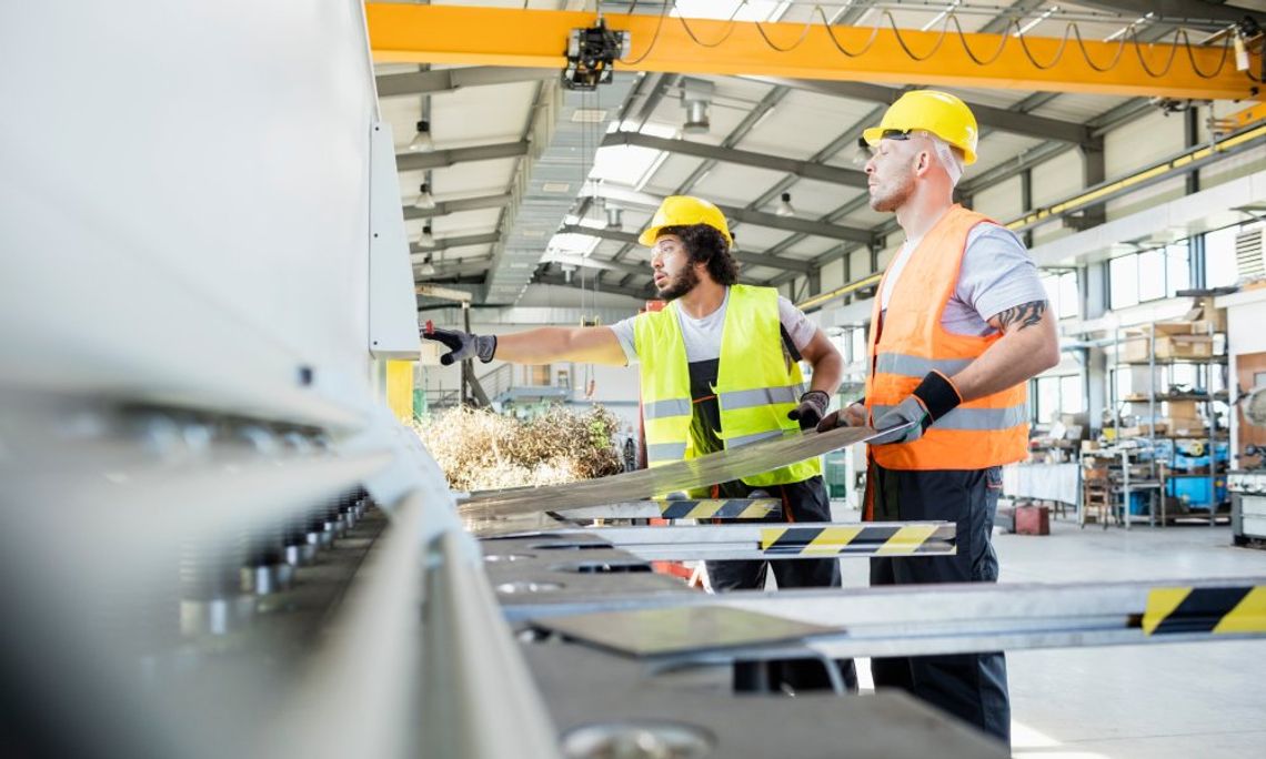 Important Materials You Need in the Manufacturing Industry Two employees wearing reflective vests in a manufacturing facility. One person is holding onto sheet metal.