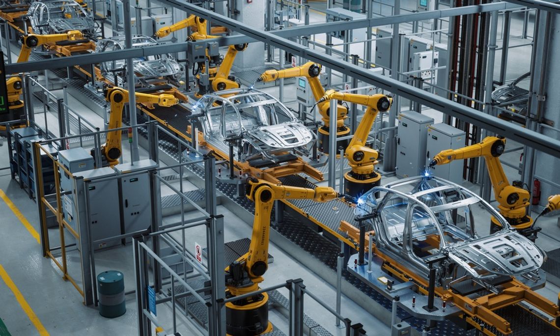 Overhead view of a factory where vehicle frames on a conveyor belt are being assembled by robotic arms.