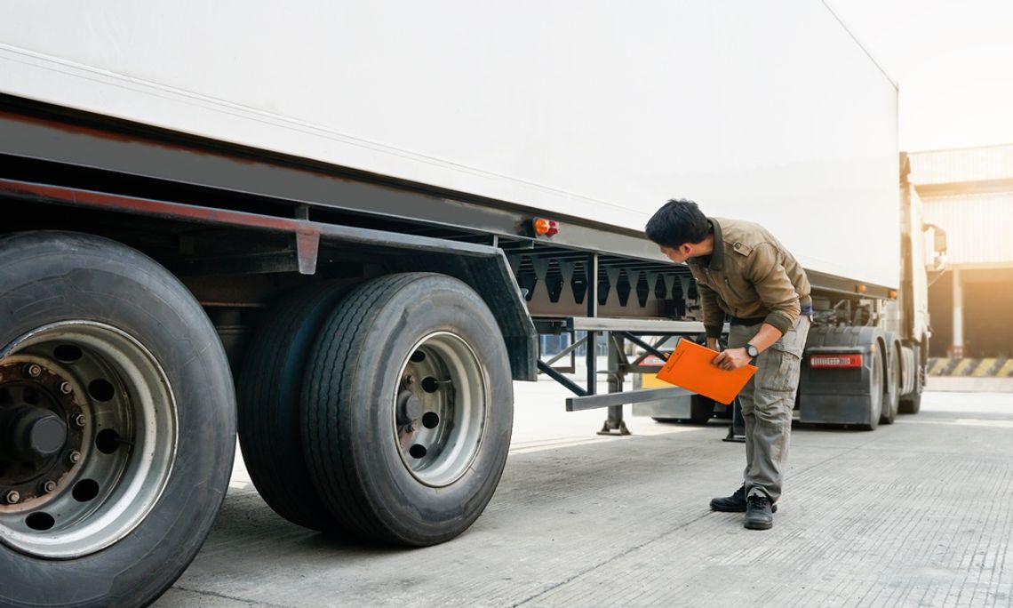 A man holding a clipboard leaning down to inspect the undercarriage of a semi-truck and its attached trailer.