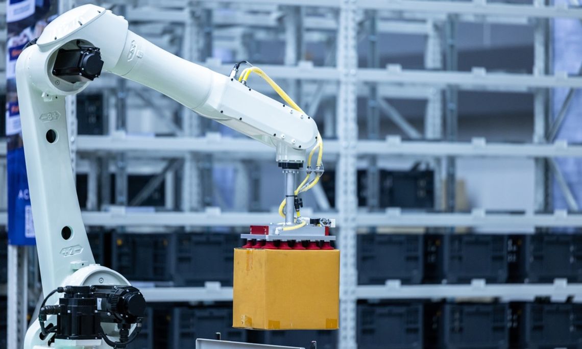 A white and black robotic arm lifts a square cardboard box in the air inside a large warehouse facility.