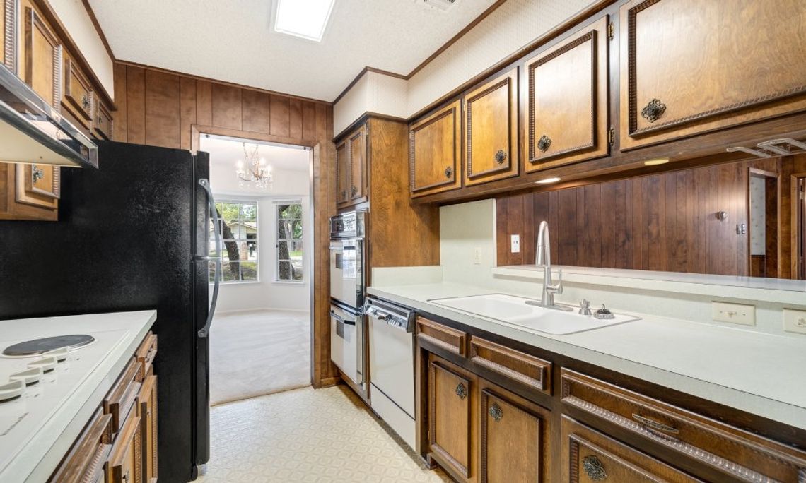 Inexpensive Ideas To Modernize Your Older Home An outdated galley kitchen contains dark-wood cabinets, a black refrigerator, white countertops, and a white wall oven.