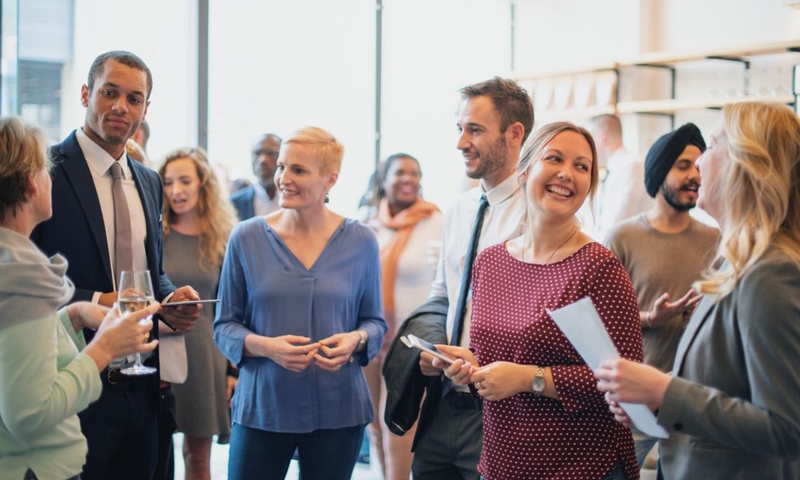 A group of professionals chatting and smiling at a corporate networking event, holding drinks and papers in a bright venue.