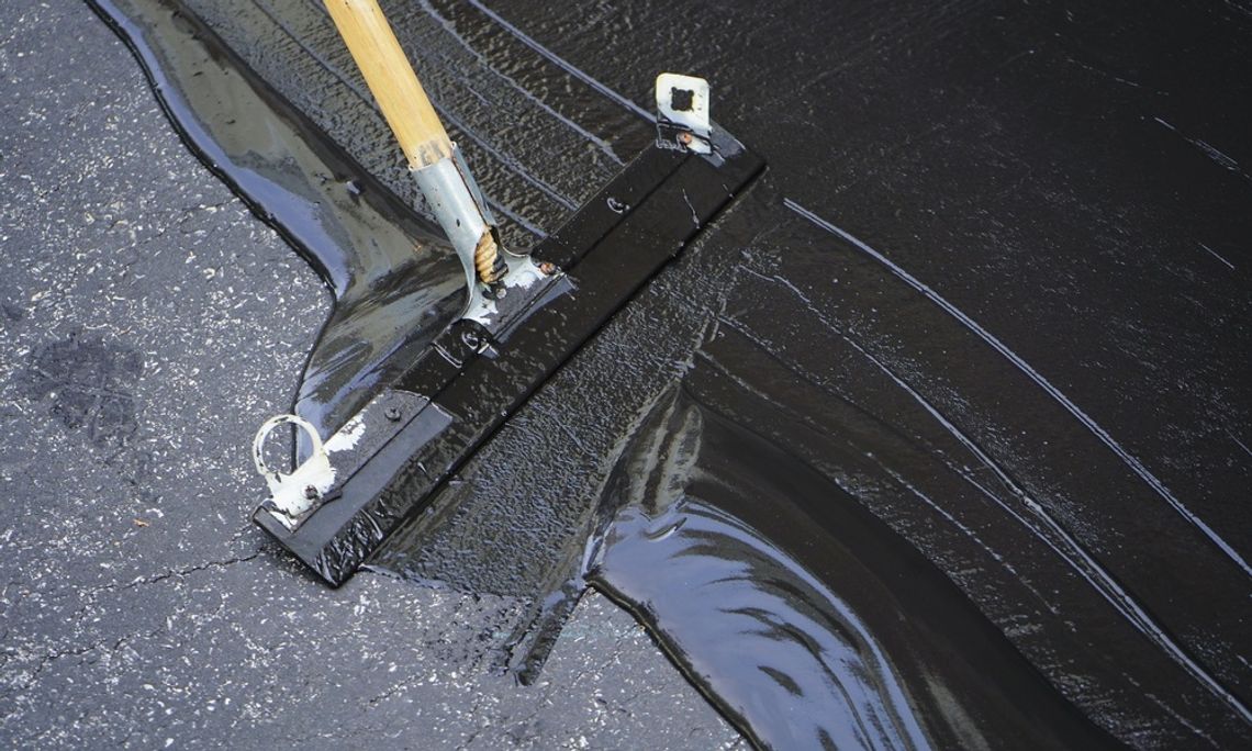 A metal squeegee with a wooden handle spreading glossy black sealant across cracked asphalt pavement.